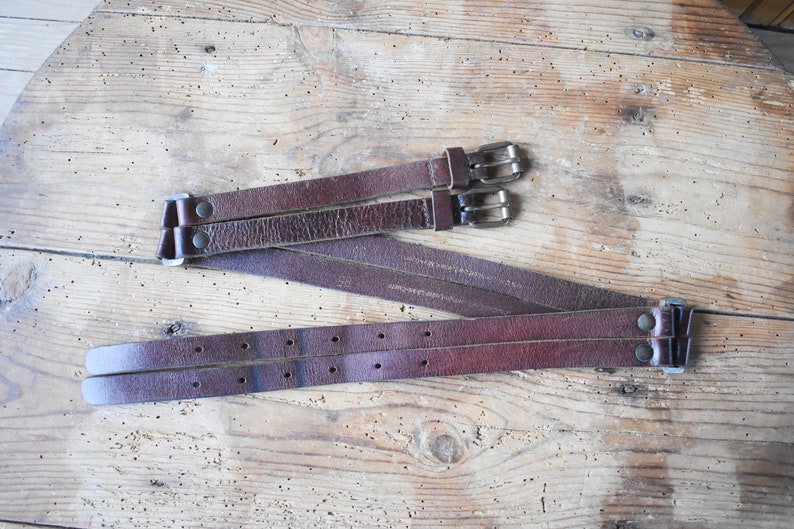 May include: Three brown leather belts with silver buckles. The belts are arranged in a criss-cross pattern on a wooden surface.