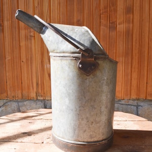 Pode incluir: Um regador vintage de metal galvanizado com um bico e uma alça. O regador tem um aspecto desgastado e rústico e está sentado em uma superfície de madeira.