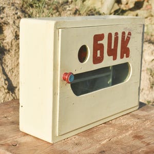 May include: An aged, cream-coloured wooden box featuring red Cyrillic lettering. The box has a circular aperture and a rectangular viewing window. A red and blue knob is on the door. It rests on a wooden surface.
