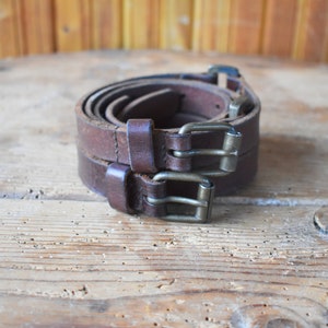 May include: Two brown leather belts with silver buckles are rolled up and stacked on top of each other on a wooden surface.