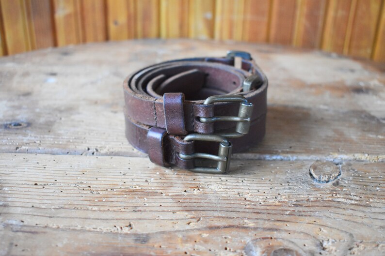 May include: Two brown leather belts with silver buckles are rolled up and stacked on top of each other on a wooden surface.