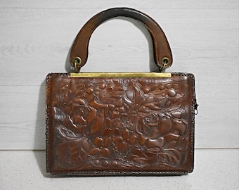 May include: A brown leather handbag with a tooled floral design and a brown leather handle. The bag has a brass clasp and a gold-toned metal frame.
