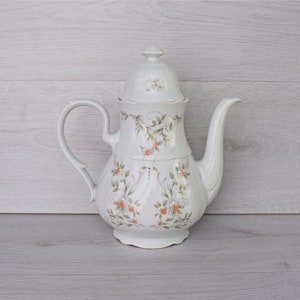 Puede incluir: Cafetera de porcelana blanca con tapa, con un diseño floral de rosas color melocotón y hojas verdes. La cafetera tiene un asa curva y un pico largo. Una pieza decorativa para servir café o té.