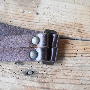 May include: Close-up of a brown leather strap with two metal rivets and a metal buckle. The strap is folded over itself and the buckle is closed.