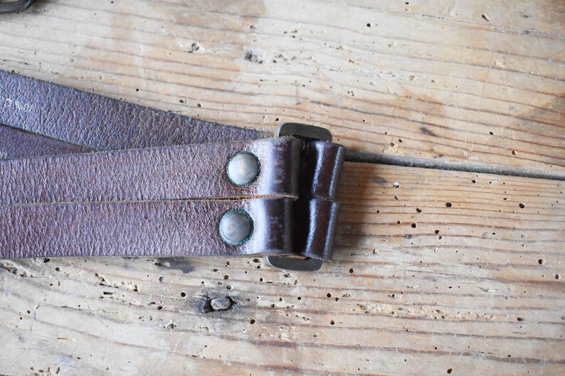 May include: Close-up of a brown leather strap with two metal rivets and a metal buckle. The strap is folded over itself and the buckle is closed.