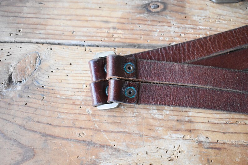 May include: Close-up of a brown leather strap with two silver buckles and turquoise accents. The strap is folded in half and the buckles are closed.