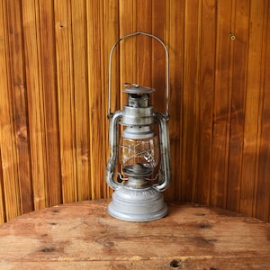 Könnte beinhalten: Eine antike silberne Petroleumlampe mit einem Glaskolben steht auf einem Holztisch. Die Lampe hat einen Griff und einen Metallfuß.