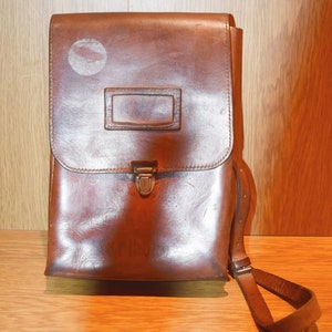 May include: A brown leather satchel with a metal clasp and a strap. The bag has a worn look and a label on the front.