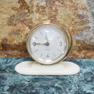 May include: A gold-colored alarm clock with a white face and black hands. The clock has a white base and says "Slavia 11 Jewels" on the face.