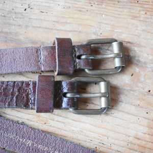 May include: Two brown leather belts with silver buckles. The belts are laying on a wooden surface.