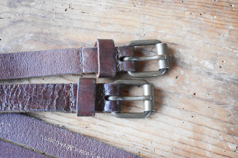 May include: Two brown leather belts with silver buckles. The belts are laying on a wooden surface.
