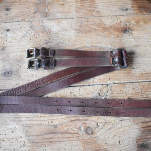 May include: Three brown leather straps with silver buckles. The straps are folded and laid out on a wooden surface.
