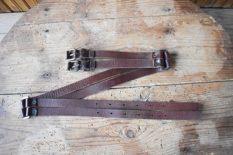 May include: Three brown leather straps with silver buckles. The straps are folded and laid out on a wooden surface.