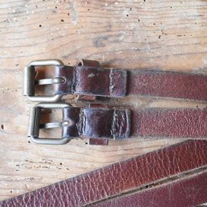 May include: Two brown leather straps with silver buckles. The straps are lying on a wooden surface.