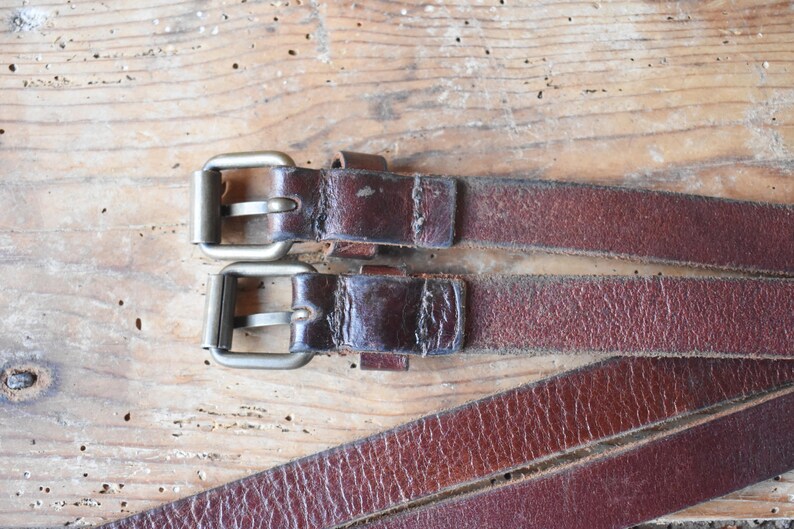 May include: Two brown leather straps with silver buckles. The straps are lying on a wooden surface.