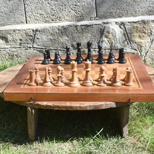 May include: A wooden chessboard with black and white squares, set up with black and white chess pieces. The board is sitting on a wooden table with a round top and three legs. The table is on a grassy surface.