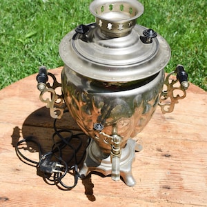 Può includere: Un samovar d'argento vintage con manici ornati e un rubinetto, appoggiato su una superficie di legno. Il samovar ha una parte superiore decorativa e un cavo di alimentazione, suggerendo che sia elettrico. Lo sfondo è erba verde.