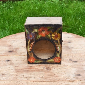 May include: A wooden box with a painted floral design. The box is square shaped and has a hole in the front. The flowers are painted in shades of red, orange, yellow, purple, and green.