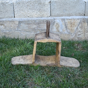 Puede incluir: Una herramienta de madera con un marco rectangular y una base plana. El marco tiene una serie de púas de madera verticales que sobresalen de la parte superior.