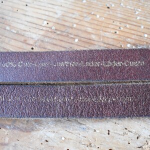 May include: Close-up of a brown leather strap with the text "100%  Genuine Leather - Sticker-Queen" embossed in gold.