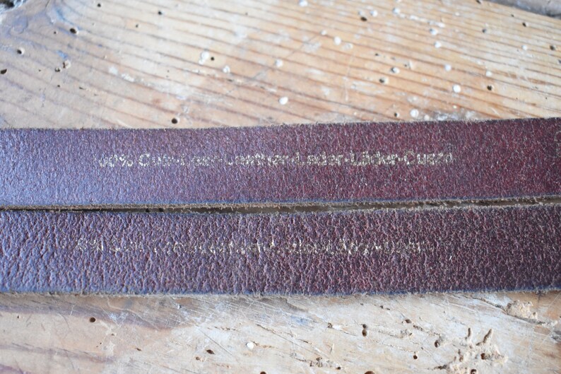 May include: Close-up of a brown leather strap with the text "100%  Genuine Leather - Sticker-Queen" embossed in gold.