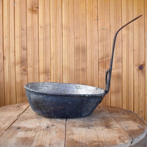May include: A vintage, black metal pan with a long handle. The pan has a silver interior and is sitting on a wooden surface.