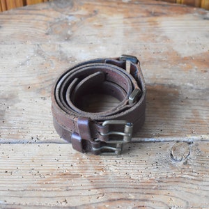 May include: Two brown leather belts with silver buckles are rolled up and placed on a wooden surface.