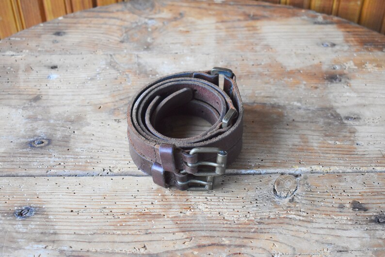 May include: Two brown leather belts with silver buckles are rolled up and placed on a wooden surface.