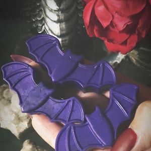 May include: Three purple bat-shaped wax melts. The wax melts are in the shape of bats with wings and a body. The wax melts are in a hand.
