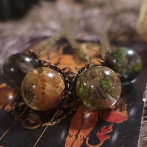 Könnte beinhalten: Eine Halskette mit fünf runden Glasperlen. Die Perlen sind mit einer Mischung aus grünen, braunen und goldenen Materialien gefüllt. Die Perlen sind an einer Bronzekette befestigt.