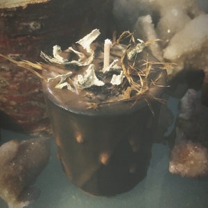 Puede incluir: Una vela marrón con una mecha de madera y hierbas secas en la parte superior. La vela está en un tarro de vidrio marrón con una superficie texturizada.