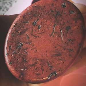 Può includere: Primo piano di un porta sapone rotondo rosso testurizzato con macchie nere. Il porta sapone è su uno stampo in silicone rosa.