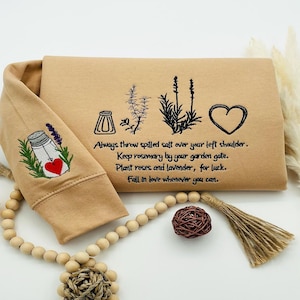 May include: Tan sweatshirt with embroidered text and images. The text reads: "Always throw spilled salt over your left shoulder. Keep rosemary by your garden gate. Plant roses and lavender, for luck. Fall in love whenever you can."