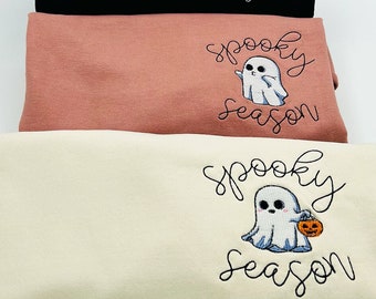 May include: Three folded sweatshirts in black, dusty rose, and cream. Each features an embroidered ghost design with the words "spooky season." The black sweatshirt has a ghost holding a sign. The cream sweatshirt has a ghost holding a pumpkin.