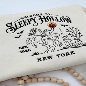May include: A cream-colored sweatshirt featuring a black screen-printed design that reads Welcome to Sleepy Hollow, Est. 1626, New York. The graphic depicts a headless horseman riding a horse, holding a flaming orange pumpkin, with a church and bats.