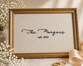 May include: A rectangular gold-toned ornate frame containing a light beige fabric insert. The fabric features black embroidered text that reads The. Morgans est. 2026. Two gold wedding bands are positioned in front of the frame on a white surface.