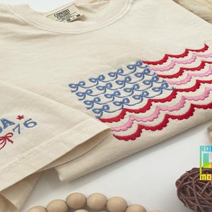 Pode incluir: Uma camiseta branca com um design de bandeira americana bordada. A bandeira é feita de laços azuis e bordas festoneadas vermelhas e rosadas. A camiseta também tem o texto "USA * 1776" bordado na manga.