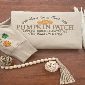 May include: Beige sweatshirt with embroidered text reading "Pumpkin Patch" and fall-themed graphics. The sleeve features embroidered leaves in yellow, green, and orange. A wooden bead garland and decorative elements are arranged on a wooden surface.