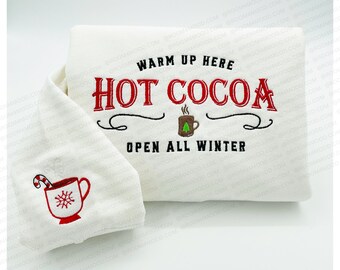 May include: A white, folded blanket and a matching pillow with embroidered text and images. The blanket features the words "Warm Up Here Hot Cocoa Open All Winter" in red and black. The pillow has a red mug with a candy cane and snowflake.
