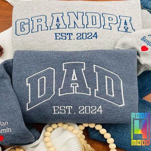 May include: Two blue and gray sweatshirts with white embroidered text. The top sweatshirt says "Grandpa Est. 2024" and the bottom sweatshirt says "Dad Est. 2024". Both sweatshirts have a small red heart with the name "Adrian Benjamin" embroidered on the sleeve.