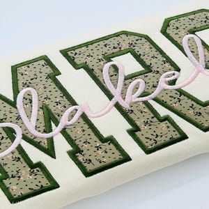 May include: A cream-coloured sweatshirt featuring large block letters with a sage green floral pattern and dark green borders. The name Blakeley is embroidered across the letters in a light pink cursive script with a raised, textured finish.