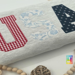Puede incluir: Un artículo de tela gris claro con las letras "USA" aplicadas. La "U" es de cuadros rojos y blancos, la "S" tiene un estampado floral azul y la "A" es azul marino con detalles florales blancos.