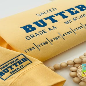 May include: Yellow sweatshirt with the text "SALTED BUTTER GRADE AA" and "NET WT. 4OZ. (113.4g)" printed on the front. The text "SWEET CREAM BUTTER GRADE AA" is printed on the sleeve.