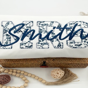Puede incluir: Cojín rectangular blanco con la palabra "Smith" bordada en azul marino. Las letras están delineadas con un estampado floral. El cojín descansa sobre una cesta de mimbre.