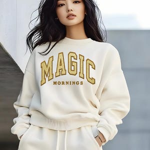 May include: Cream-colored sweatshirt and matching sweatpants. The sweatshirt features the text "MAGIC MORNINGS" in gold block letters. The model is wearing a bracelet and has long, dark hair.