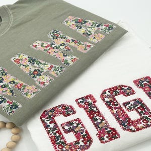 Könnte beinhalten: Zwei T-Shirts, eines in Salbeigrün und eines in Weiß, werden gezeigt. Das salbeigrüne T-Shirt hat das Wort "MAMA" in Buchstaben aus Blumenstoff. Das weiße T-Shirt zeigt das Wort "GIGI" in Buchstaben aus Blumenstoff. Beide sind gefaltet.