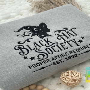 May include: A light gray sweatshirt with the embroidered text "Black Hat Society" in black. The design includes a witch silhouette, stars, and the phrase "Proper Attire Required Est. 1692". The sweatshirt is folded on a white surface.