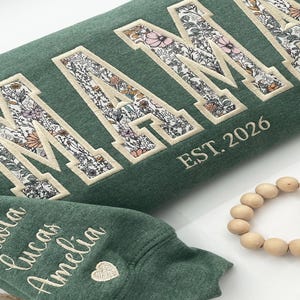 May include: Green sweatshirt with "MAMA" embroidered in large, floral-filled letters. The text "EST. 2026" is below the main text. The sleeve features embroidered names and a heart.