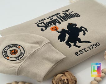 May include: Beige sweatshirt and cuff featuring a black silhouette of the Headless Horseman and the text "The Legend of Sleepy Hollow" with an orange pumpkin. The sweatshirt also has "EST. 1790". A matching cuff has a circular patch with a pumpkin.