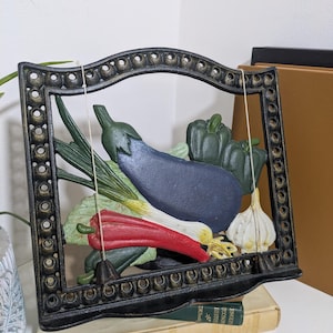 May include: A black cast iron cookbook stand featuring a decorative frame with circular cutouts. The centre displays a relief of colourful vegetables including a purple aubergine, a red chilli pepper, green bell peppers, and a white garlic bulb.